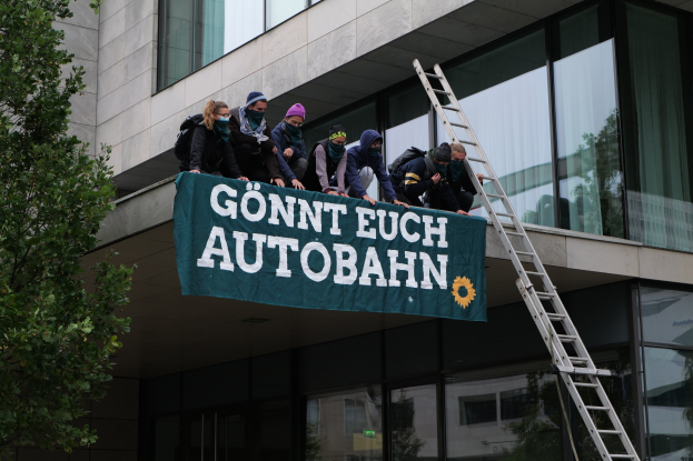 Eine Gruppe von Menschen auf dem Dach eines Gebäudes stehend, die eine Fahne halten, mit einem Baum links und Glasfenstern im Hintergrund, wahrscheinlich protestierend im Support der deutschen Regierung.