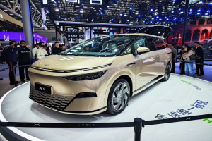 Das Xpeng X9-Auto auf einer Automesse, umgeben von Menschen, mit Pfählen, einem Zaun, einem Anzeigebildschirm, einem Gebäude und einem Dach mit Deckenbeleuchtung im Hintergrund.