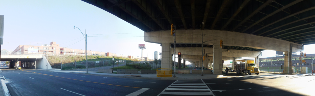 Ein Lastwagen fährt unter einer Brücke auf einer Stadtstraße mit Gebäuden, Straßeninfrastruktur und Fahrzeugen unter einem klaren blauen Himmel.
