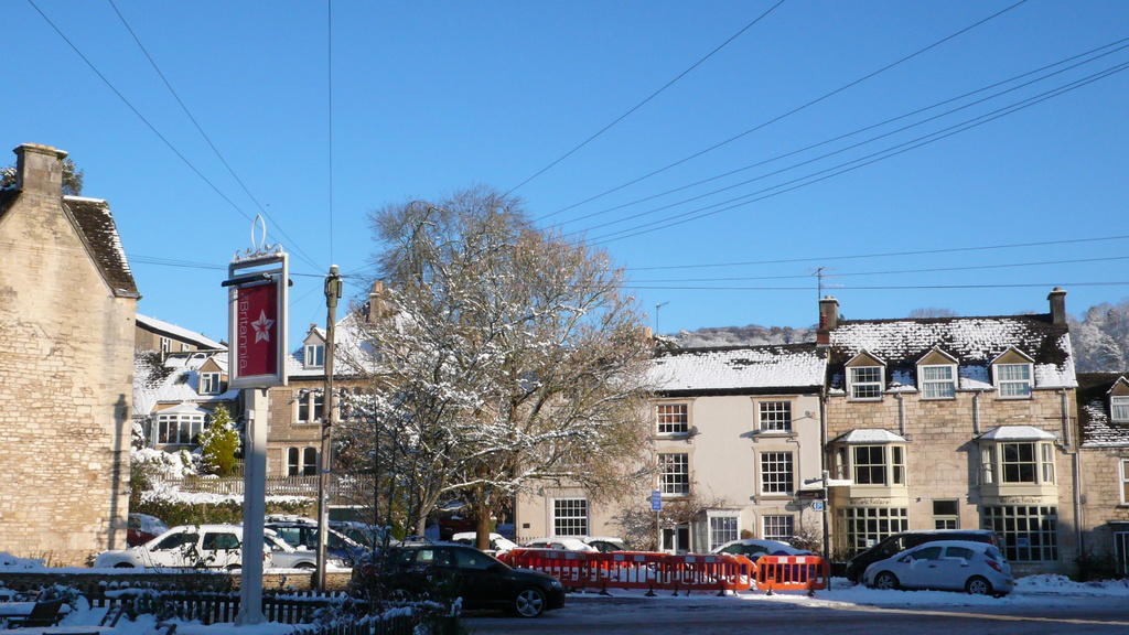 Fahrzeuge auf einer schneebedeckten Straße mit Häusern, Bäumen und Strommasten im Hintergrund, alles mit Schnee bedeckt.