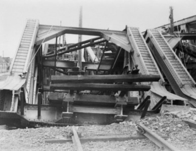 Schwarzes und weißes Foto einer Bahnschiene, die zu einer zerfallenden Brücke mit kaputten Geländern und Schutt führt, umgeben von Gebäuden unter einem klaren Himmel.