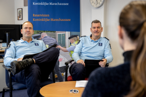 Zwei Polizeibeamte sitzen an einem Tisch mit einer Frau, umgeben von Dokumenten und Gegenständen, mit einem Wandbanner, einer Uhr und Fotorahmen im Hintergrund und einem zweiten Tisch mit einem Computer-Setup links.