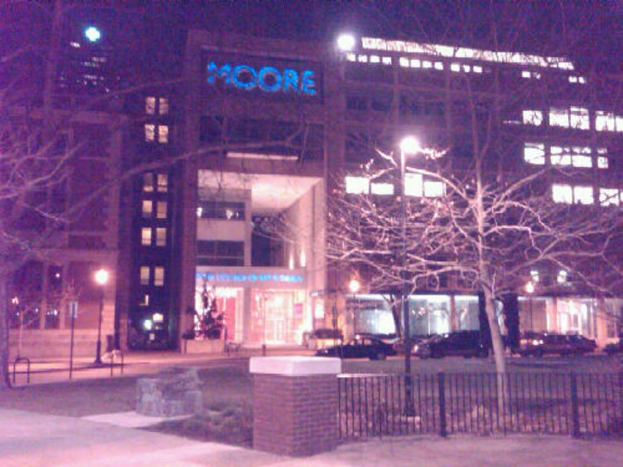 Ein krankenhausähnliches Gebäude mit Fahrzeugen, Bäumen und einem Zaun davor.