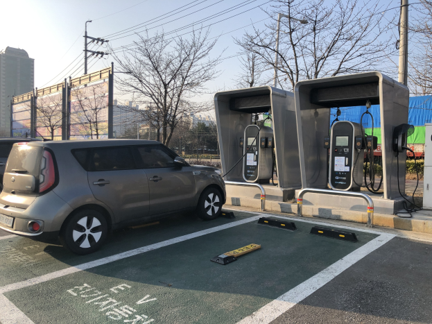 Elektrisches Auto-Ladestation auf einem Parkplatz mit Bäumen, Gebäuden, Strommasten und einem klaren Himmel.