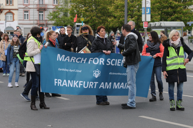Eine vielseitige Gruppe von Menschen marschiert die Straße entlang, hält ein "March for Science Frankfurt am Main"-Schild hoch, mit Bäumen, Gebäuden und einem klaren Himmel im Hintergrund.