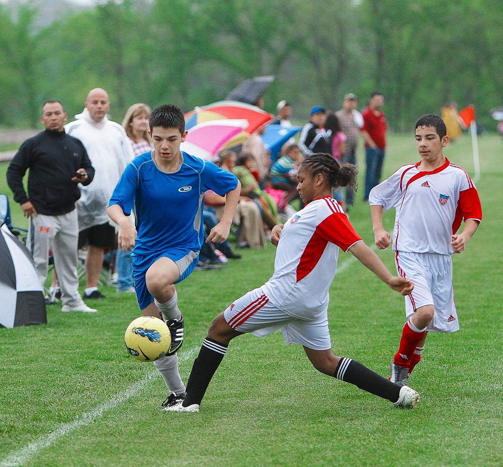 Drei Personen spielen Fußball mit einem gelben Ball, während eine Gruppe von Menschen hinter ihnen steht und sitzt und Regenschirme hält, mit Bäumen im Hintergrund.