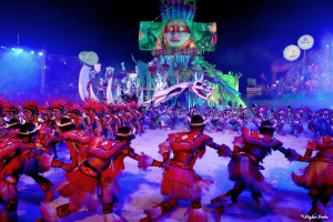 Eine Gruppe von Menschen in farbenfrohen Kostümen tanzt auf einer Bühne vor einem sitzenden Publikum, mit Lichtern, Pfählen, Ballons und einem Bildschirm, der eine Person in der Hintergrund zeigt, unter einem sichtbaren Himmel, bei der Rio-Karneval 2014.
