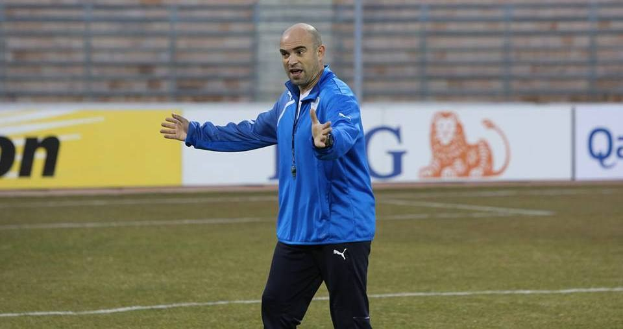 Ein Mann in einer blauen Jacke steht auf einem Fußballfeld während eines Trainings, blickt entschlossen nach vorne und ist von Tafeln mit Text und Logos umgeben.