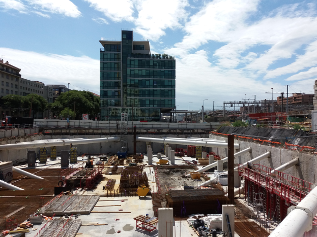 Eine Baustelle mit einem großen Gebäude im Hintergrund, umgeben von Bäumen, Pfählen, Lampen, unter einem bewölkten Himmel, das scheinbar der Sitz der Europäischen Union ist.