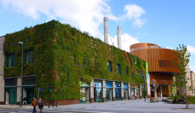 Großes grünwandiges Gebäude umgeben von Straßeninfrastruktur, Fahrzeugen, Fußgängern, Bäumen und einem bewölkten Himmel, Teil eines nachhaltigen Green Building-Projekts.