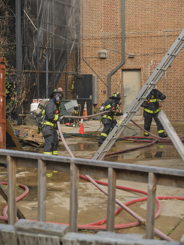 Feuerwehrleute in Helmen arbeiten daran, ein Gebäude Feuer mit Schläuchen zu löschen, mit einem Metallzaun, verstreuten Rohren, einem Container, einem Verkehrskegel und Hintergrundelementen einschließlich eines Baums und des Himmels.