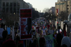 Menschen demonstrieren auf einer Straße und halten Plakate, mit Gebäuden, Bäumen, Laternen und dem Himmel im Hintergrund.