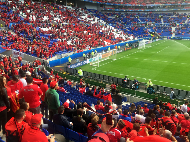 Eine große Menschenmenge in einem Stadion schaut einem Fußballspiel zu, mit Menschen auf Sitzen und Stehplätzen, Fahnen, einem Torpfosten und Deckenlichtern.