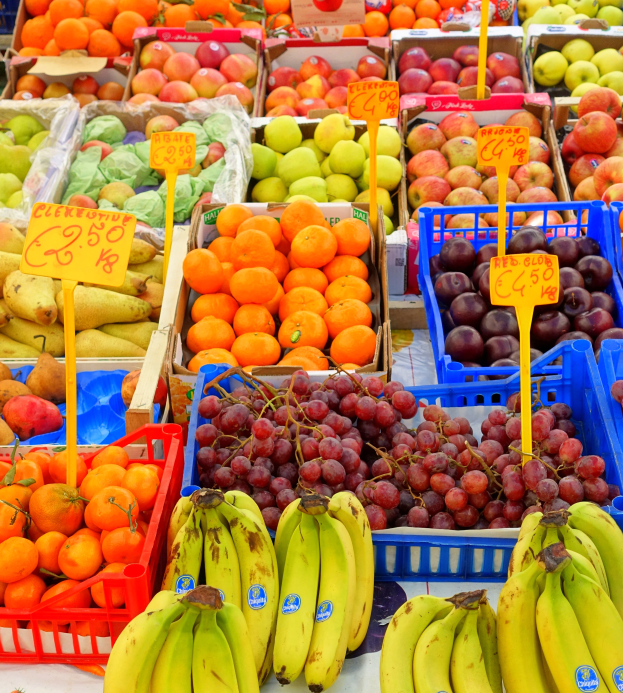 Ein Marktstand mit einer Vielfalt an Obst und Gemüse, darunter Bananen, Äpfel, Orangen und Trauben, mit Preisschildern an jedem Artikel.