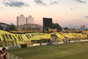 Fußballfeld mit einer Gruppe von Menschen, einem Torpfosten, einer 'Boca Juniors gegen Real Sociedad'-Tafel, Pflanzen, einem Anzeigebildschirm, Pfählen, einem Baum und einer bewölkten Himmel.