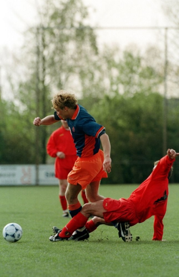 Zwei Männer beim Fußballspielen auf einem Feld mit Bäumen, Pfosten und einem klaren blauen Himmel, einer trägt Fußballschuhe während eines Spiels.