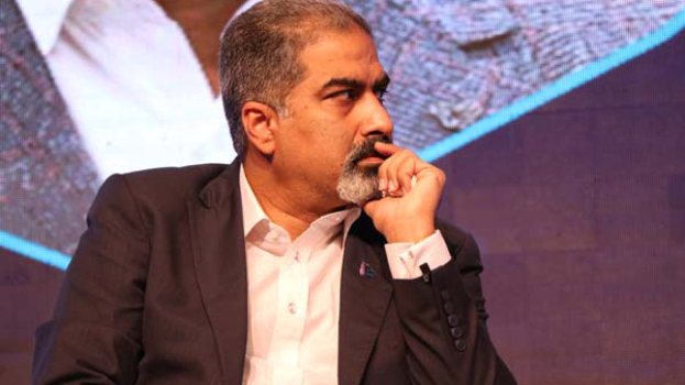 Ein Mann im Anzug und Krawatte sitzt auf einer Bühne und sieht nachdenklich auf einen Bildschirm.