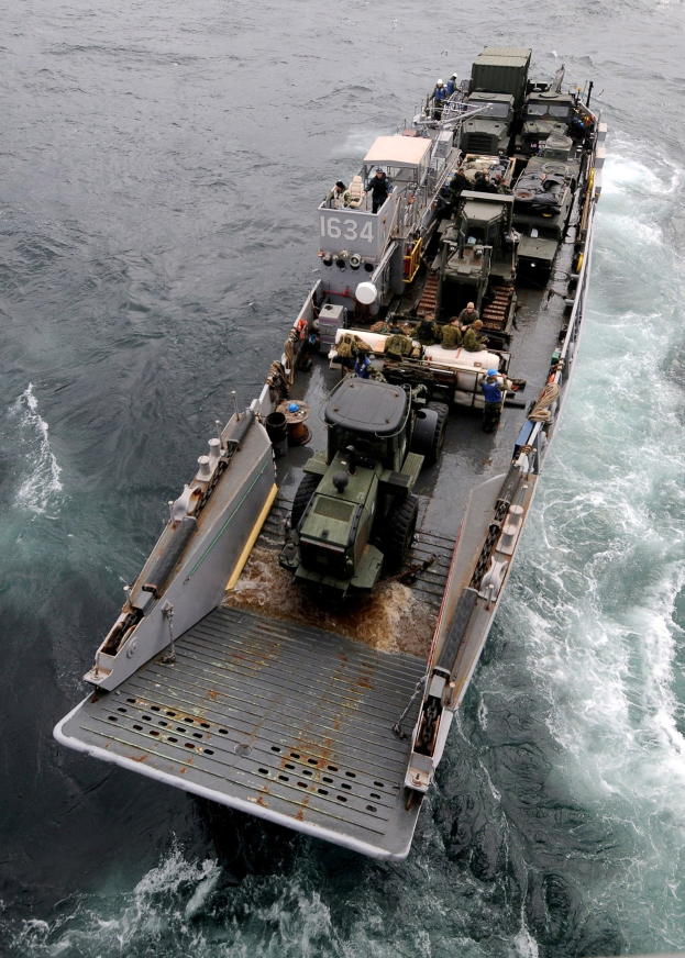 Luftaufnahme eines großen Bootes auf dem Meer mit einem Militärfahrzeug darauf, umgeben von Menschen und Gegenständen, und einem Wasserzeichen unten.
