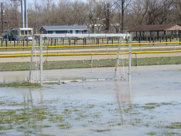 Ein Fußballtor steht in der Mitte eines überfluteten Feldes, umgeben von Gras und Wasser, mit Hütten, Pfählen, Bäumen, Fahrzeugen und einem klaren blauen Himmel im Hintergrund.