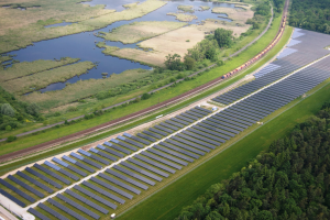 Luftaufnahme eines Solarparks mit Solarpanelen, umgeben von Bäumen, Gras, Pflanzen und Wasser, mit einem Zug auf einer nahen Bahnstrecke.