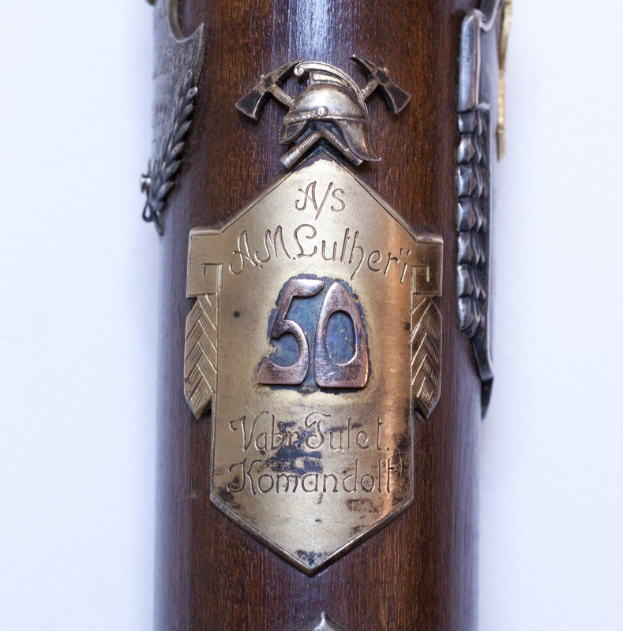 Nahaufnahme einer braunen Waffe mit einer Metallplakette, auf der '50. Jahrestag der deutschen Armee' steht, vor einem weißen Hintergrund.