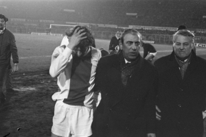 Schwarzes und weißes Bild einer Gruppe von Männern, die auf einem Fußballfeld mit einem Stadion im Hintergrund stehen und enttäuscht nach einem Sieg aussehen.