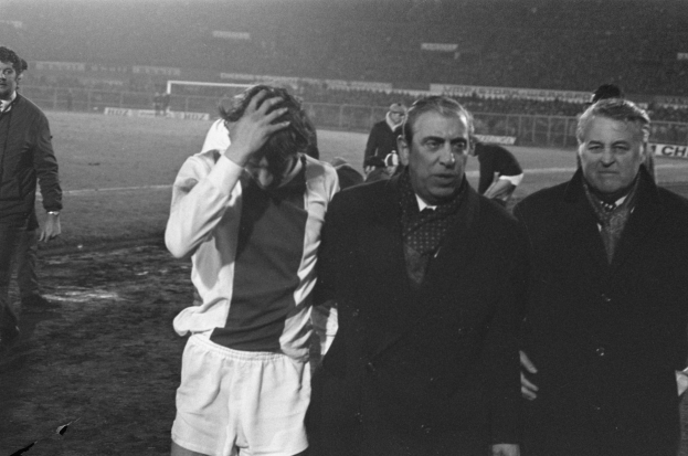 Schwarzes und weißes Bild einer Gruppe von Männern, die auf einem Fußballfeld mit einem Stadion im Hintergrund stehen und enttäuscht nach einem Sieg aussehen.