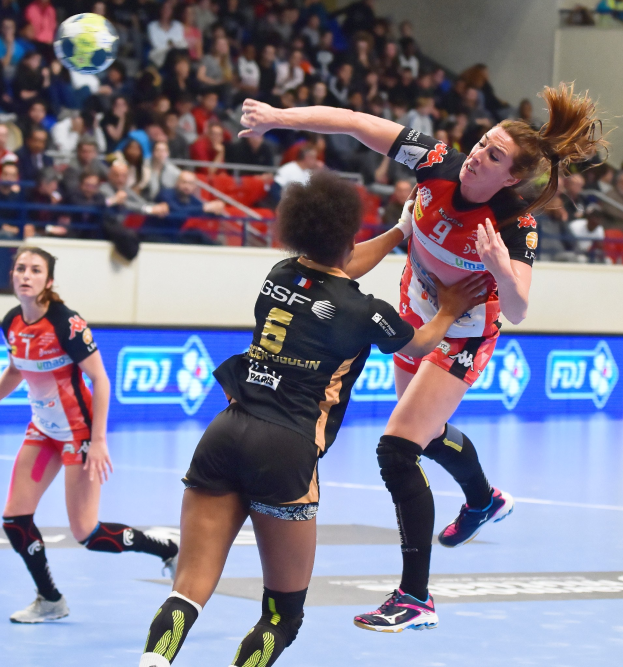 Frauen spielen Handball auf einem Feld mit einem Ball in der Luft, in einer Arena mit Zuschauern im Hintergrund.