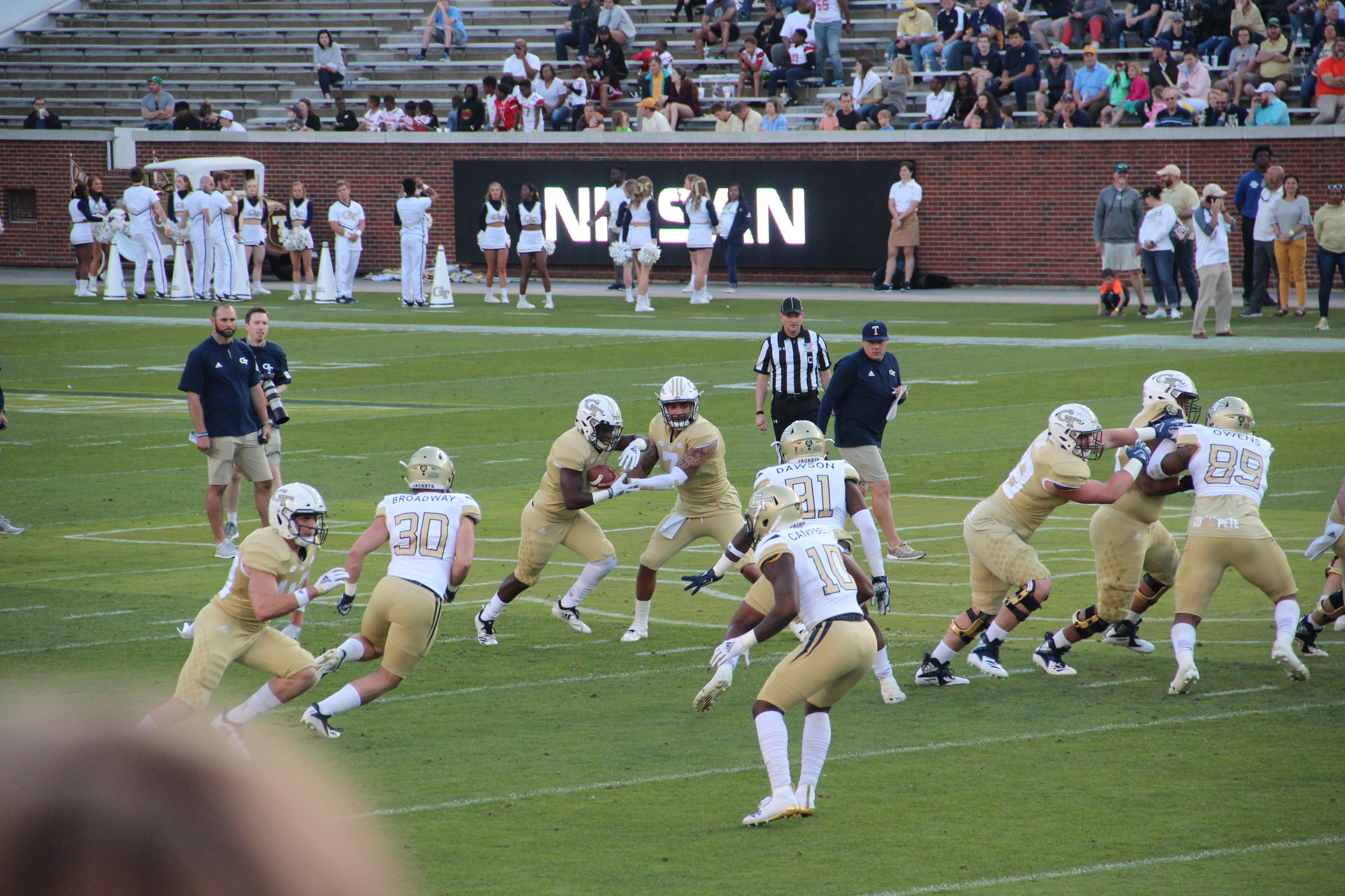 Eine Gruppe von Menschen, die Fussball auf einem Feld spielen, tragen Sportbekleidung und Helme, einige halten K├Ąmeras, mit Zuschauern auf B├Ąnken und einer Wand mit Text im Hintergrund.