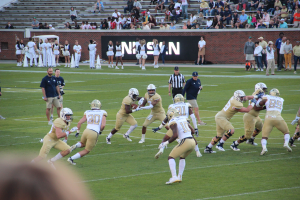 Eine Gruppe von Menschen, die Fussball auf einem Feld spielen, tragen Sportbekleidung und Helme, einige halten K├Ąmeras, mit Zuschauern auf B├Ąnken und einer Wand mit Text im Hintergrund.