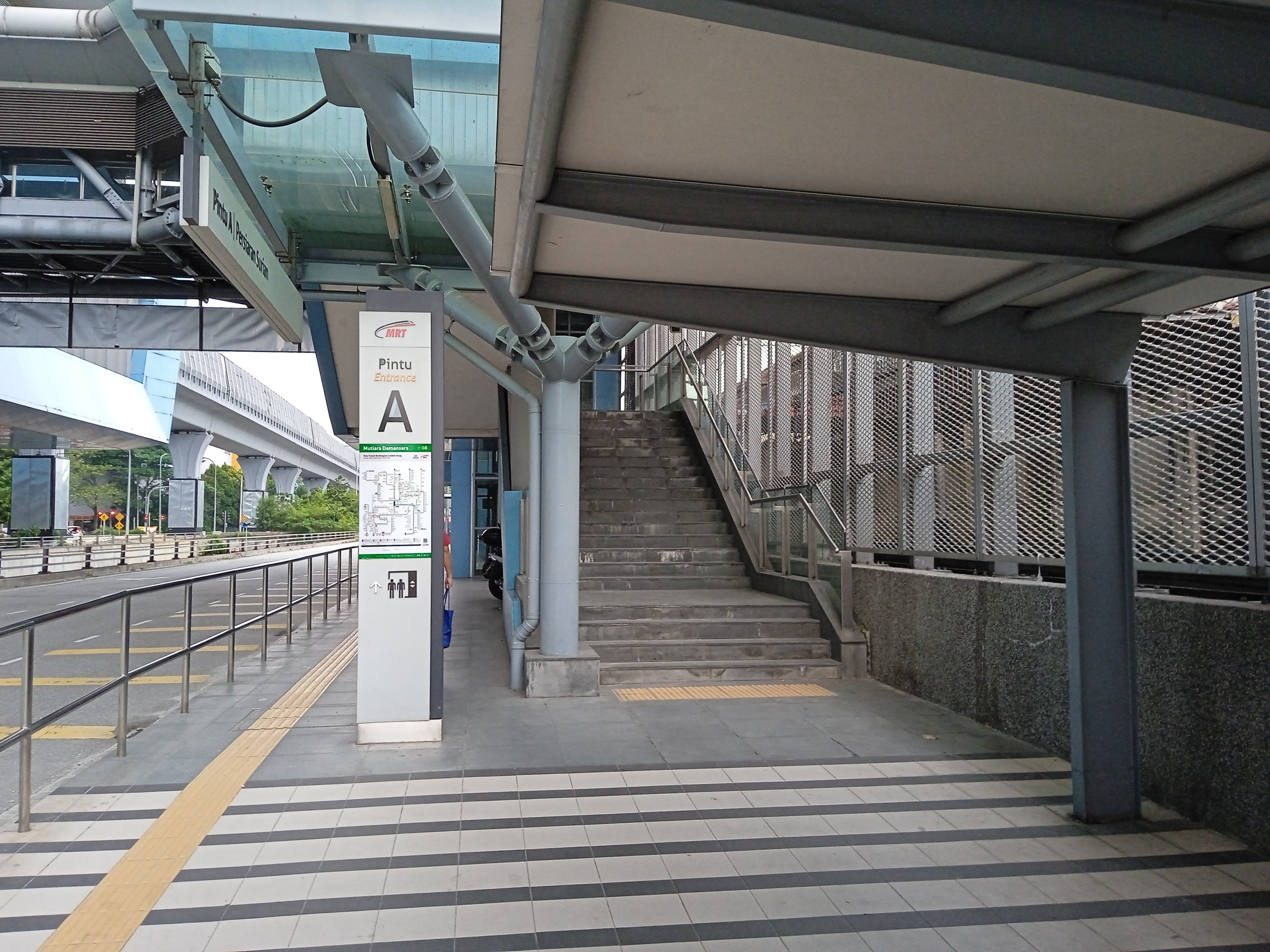 Ein Bahnsteig mit Treppe, eine Straße mit Geländer auf der linken Seite und eine Tafel mit Text oben auf dem Bahnsteig, mit Bäumen, Pfählen, Gebäuden und einer Brücke im Hintergrund.