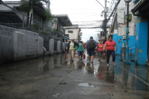 Menschen gehen eine regennasse Straße entlang, einige tragen Taschen und Schirme, mit Gebäuden, einem Metallzaun, einem Straßenpfahl, Drähten, einer Schautafel, Pflanzen und einem Baum an den Seiten.