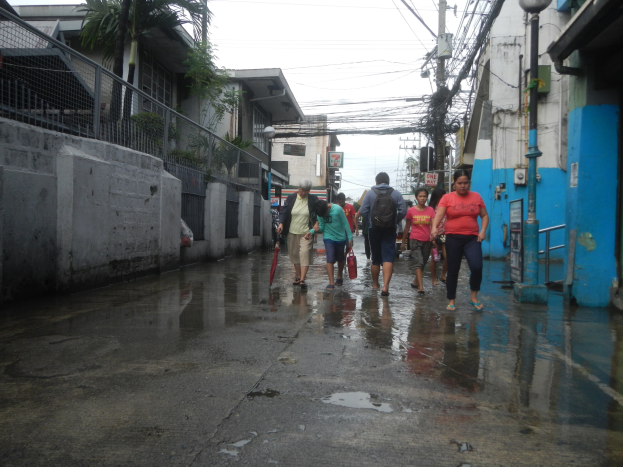 Menschen gehen eine regennasse Straße entlang, einige tragen Taschen und Schirme, mit Gebäuden, einem Metallzaun, einem Straßenpfahl, Drähten, einer Schautafel, Pflanzen und einem Baum an den Seiten.
