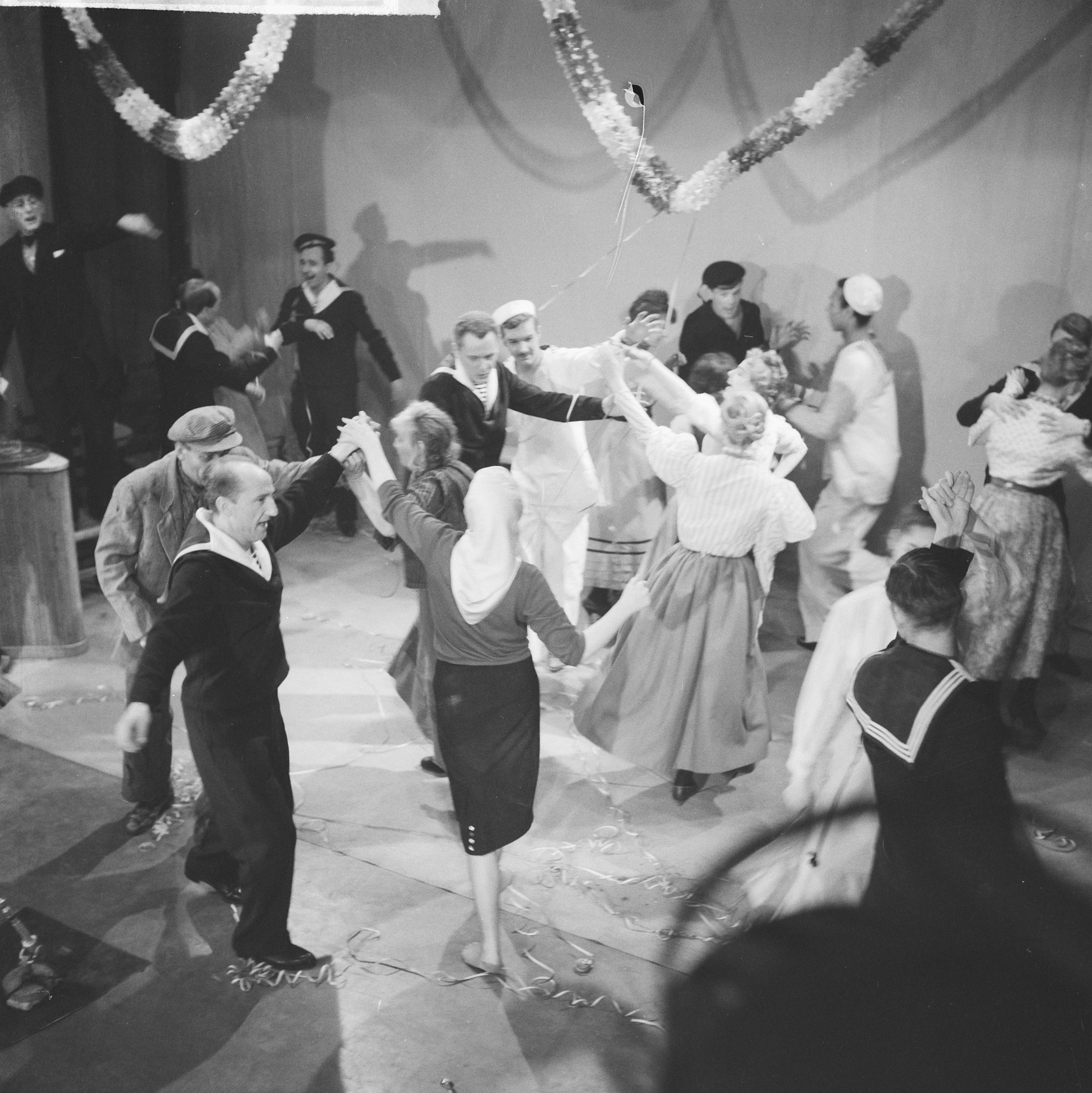 Schwarzes und weißes Bild von Menschen, die auf einer Tanzfläche tanzen, mit einer dekorierten Wand dahinter und einem Tisch mit Gegenständen links.