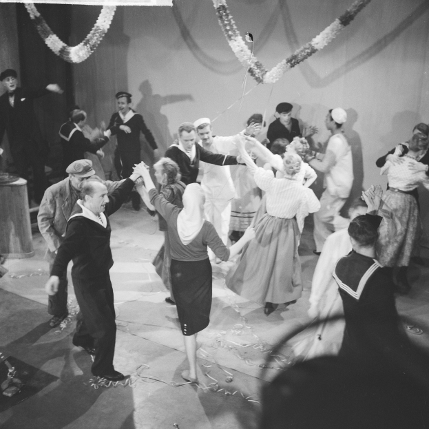Schwarzes und weißes Bild von Menschen, die auf einer Tanzfläche tanzen, mit einer dekorierten Wand dahinter und einem Tisch mit Gegenständen links.