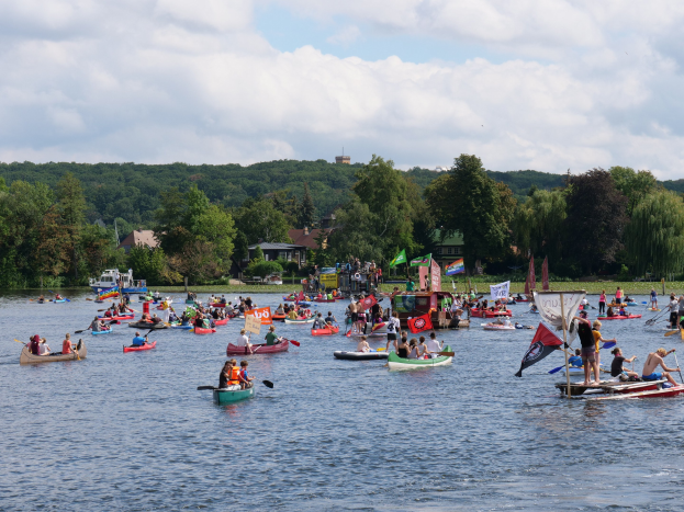 Eine große Gruppe von Menschen in Kanus auf einem Gewässer während eines Rennens, einige halten Paddel und Fahnen, mit Bäumen, Gebäuden und einem bewölkten Himmel im Hintergrund.