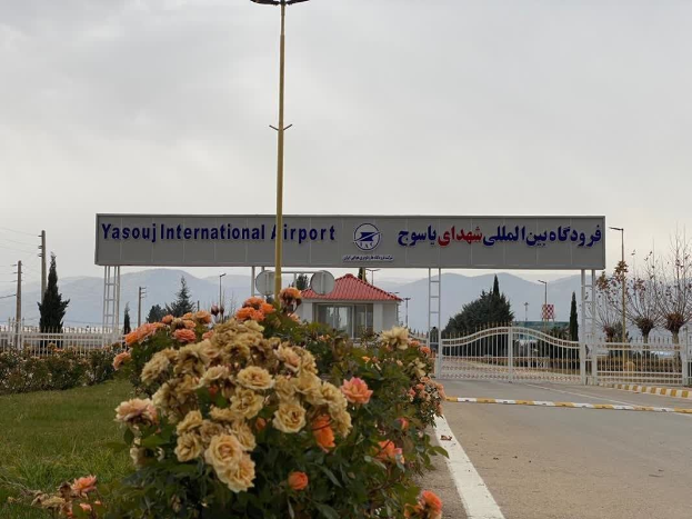 Der Eingang zum Yasouj International Airport, mit einer Straße, bepflanzten Blumen, einem Zaun, einem Haus, Bäumen, Strommasten mit Drähten, fernen Hügeln und einem bewölkten Himmel.