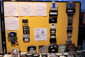 Ein Tisch voller verschiedener Festplatten, eine Person rechts stehend und ein Brett mit Papieren im Hintergrund.