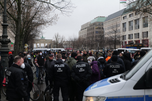 Große Gruppe von Menschen vor einem Polizeibus auf einer Straße mit Bäumen, Laternenmästen und Gebäuden, einige tragen Kappen und Masken, Fahrräder im Vordergrund, Bogen mit Säulen und Flagge im Hintergrund, Himmel über dem Bild sichtbar.