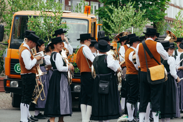 Eine Gruppe von Menschen in traditioneller bayrischer Tracht, die Musikinstrumente spielen und Taschen tragen, mit Bäumen und Gebäuden im Hintergrund und einem geparkten Fahrzeug.