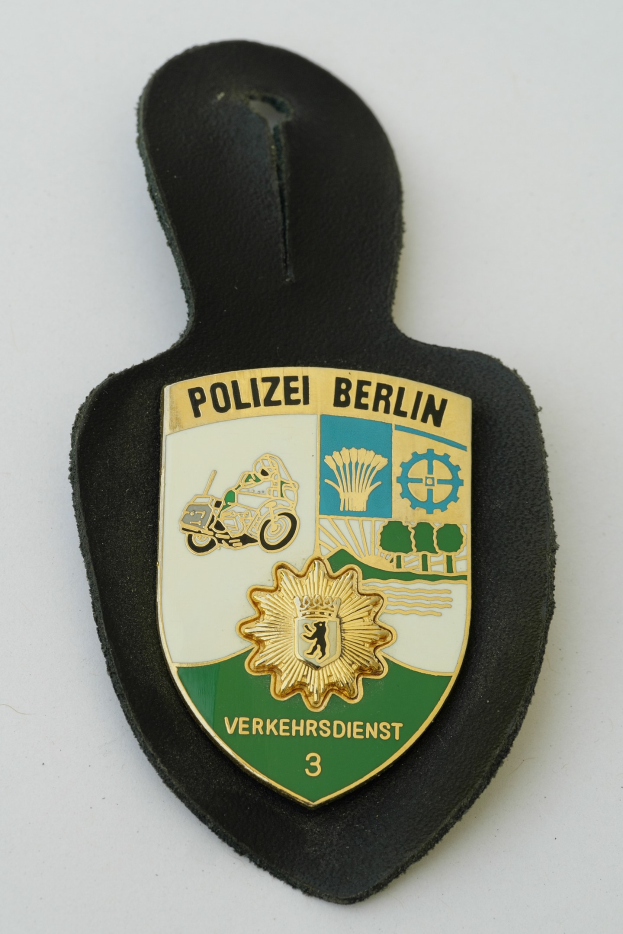 Ein schildförmiges Polizeibädge mit 'Polizei Berlin' in schwarzer Schrift, das ein weißes Stern auf einem blauen Hintergrund und einen weißen Rand hat, liegt auf einer Oberfläche.