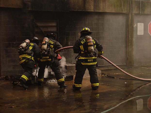 Feuerwehrleute in Helmen und Schutzausrüstung löschen ein Feuer mit einem Schlauch, während sie Sauerstoffzylinder tragen, mit Rauch im Hintergrund und einer Leitung, einem Schild, einer Wand und einer Tür in der Nähe.
