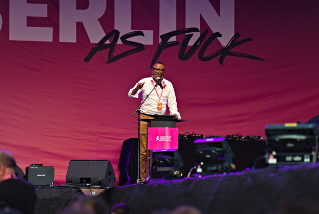 Mann in weißem Hemd und braunen Hosen, der in ein Mikrofon spricht, vor einer pinken 'Berlin as Fuck'-Fahne auf der Bühne.