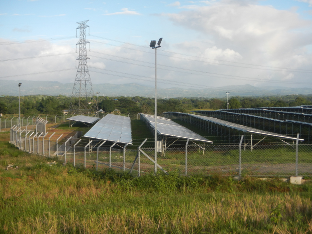 Ein Solarpanel-Feld, das von einem Zaun umgeben ist, mit Gras und Pflanzen im Vordergrund, Bäumen und Übertragungsmasten im Hintergrund und einem bewölkten Himmel.