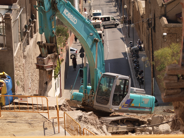 Bagger im Einsatz auf einer städtischen Baustelle mit Gebäuden, Fahrzeugen, Fußgängern und Infrastruktur im Hintergrund.