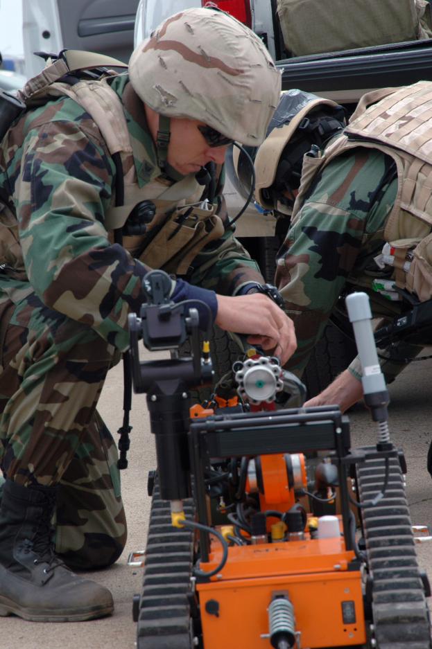 Zwei Männer in Tarnuniformen und Helmen arbeiten an einem kleinen Roboter auf dem Boden, mit Fahrzeugen und Taschen im Hintergrund.