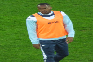 Ein Mann in einem orangen Hemd und blauen Hosen läuft über ein grünes Feld und sieht entschlossen aus.