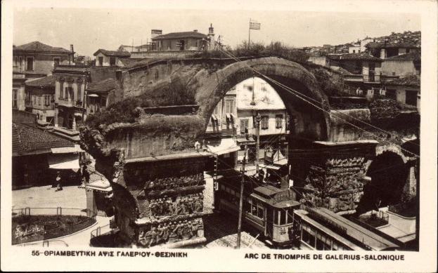 Schwarz-weiß-Foto einer Stadtstraße mit einer Bogengracht in der Mitte, umgeben von Gebäuden, Bäumen, Pfählen, Drähten und Menschen, mit Text unten.