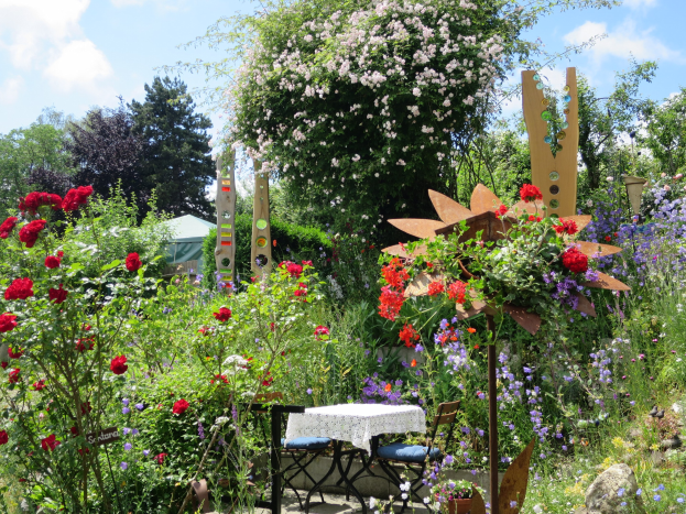 Ein Garten mit bunten Blumen und Pflanzen, mit einem Tisch mit einer Decke und Stühlen, vor Bäumen und einem bewölkten Himmel auf der Chelsea Flower Show.