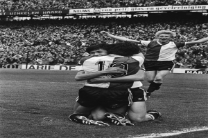 Drei Fußballspieler feiern auf dem Boden ein Tor vor einer jubelnden Menge in einem schwarz-weißen Stadion.